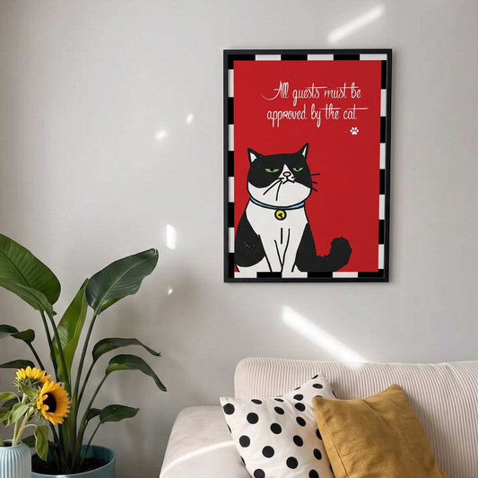 A framed wall art print featuring a black-and-white cat on a red background with the text 'All guests must be approved by the cat,' styled above a sofa with pillows in a sunlit living room.