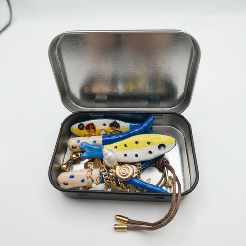 An open silver metal tin containing several handmade ceramic fish-shaped jewelry pieces in blue, yellow, white, and beige, with gold-tone charms and a brown cord tassel, on a white background.