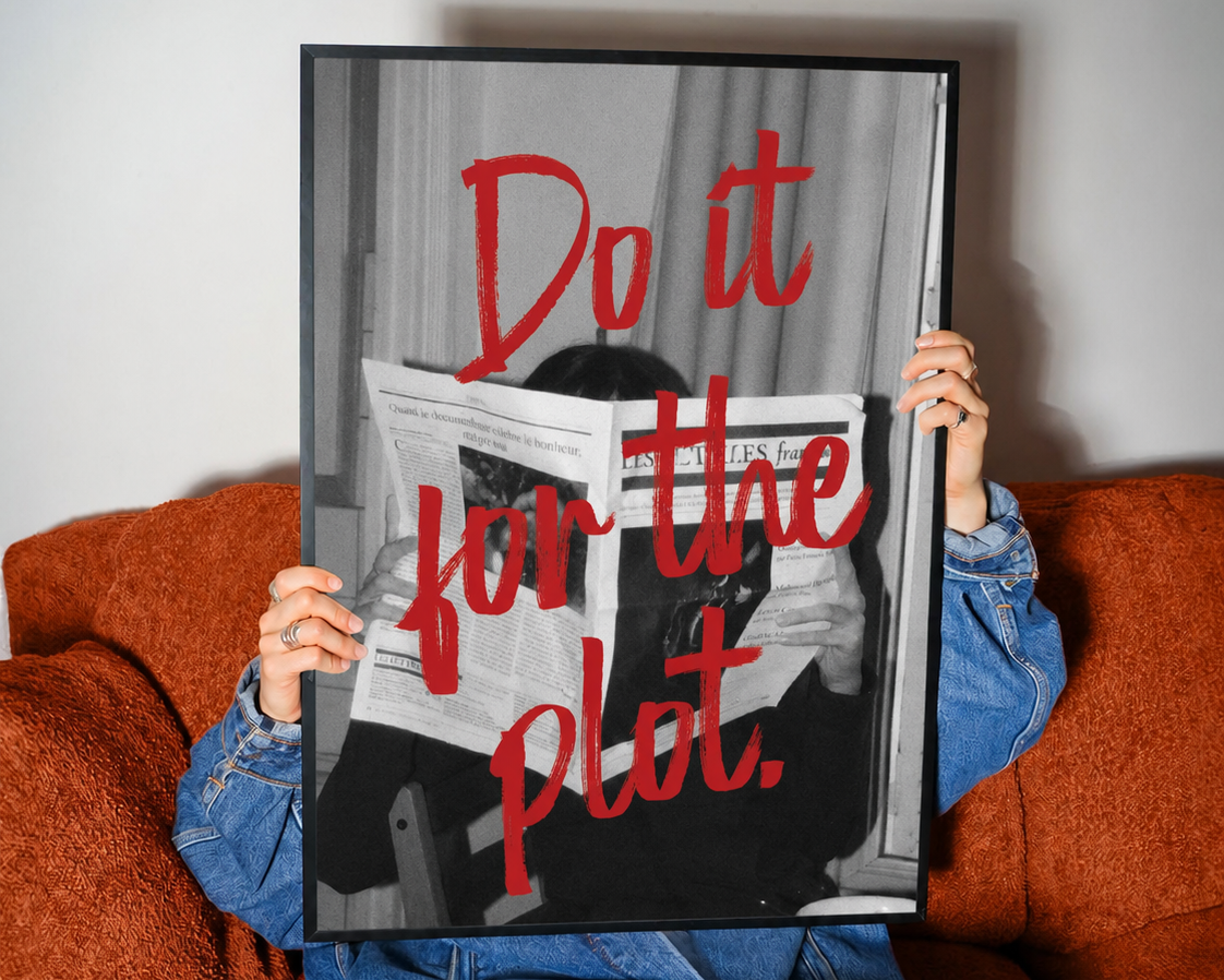 A person sitting on an orange-brown couch holds up a framed black-and-white art print with bold red text reading 'Do it for the plot.' The print features a person reading a newspaper, and the person holding it wears a blue denim jacket and jeans.