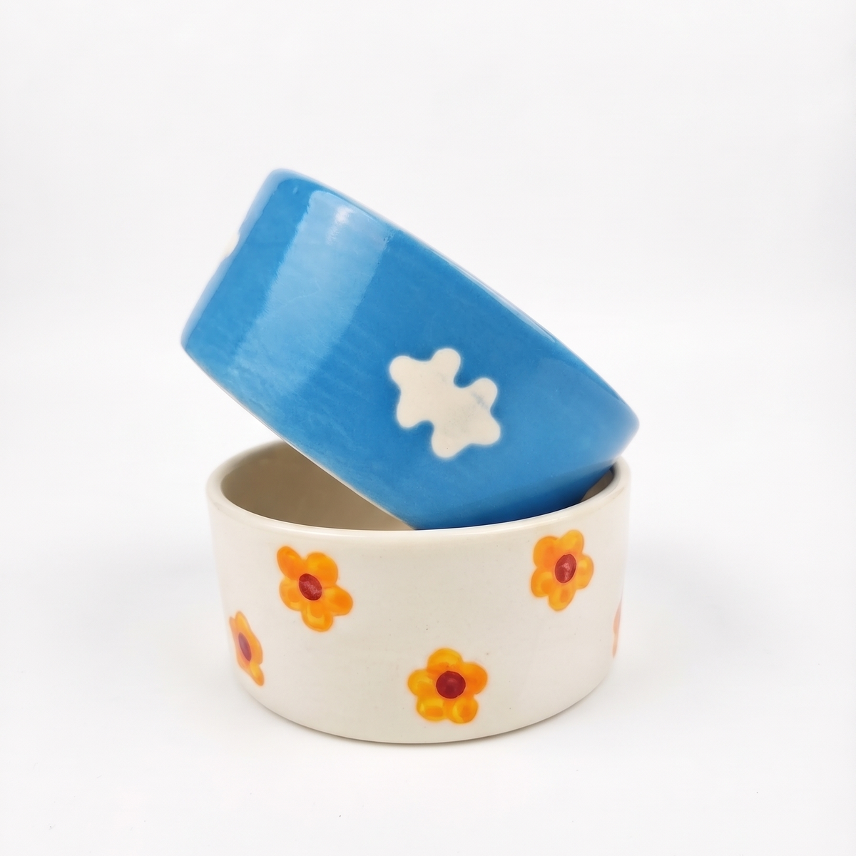 Two ceramic bowls stacked on a white background. The bottom bowl is off-white with orange and yellow flower patterns, and the top bowl is bright blue with a white flower pattern.