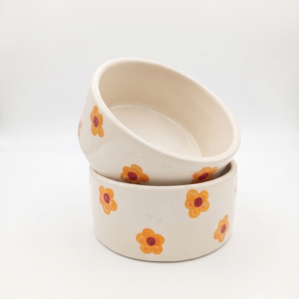 Two stacked ceramic bowls with a warm cream base and a pattern of small orange flowers with dark red centers, set against a seamless white background.