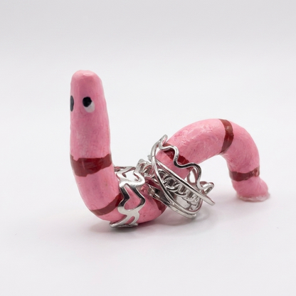Pink worm-shaped ring holder with silver heart and anchor charms on a white background