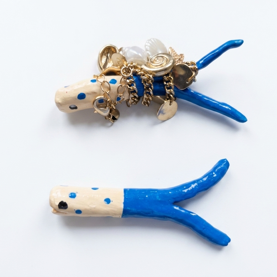 Two decorative fish shaped necklace holder on a white background. One holder is cream with blue polka dots and topped with gold-tone charms, and the other is cream and blue with a small black eye.
