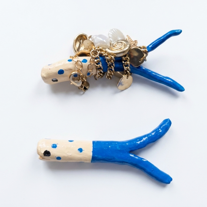 Two decorative fish shaped necklace holder on a white background. One holder is cream with blue polka dots and topped with gold-tone charms, and the other is cream and blue with a small black eye.