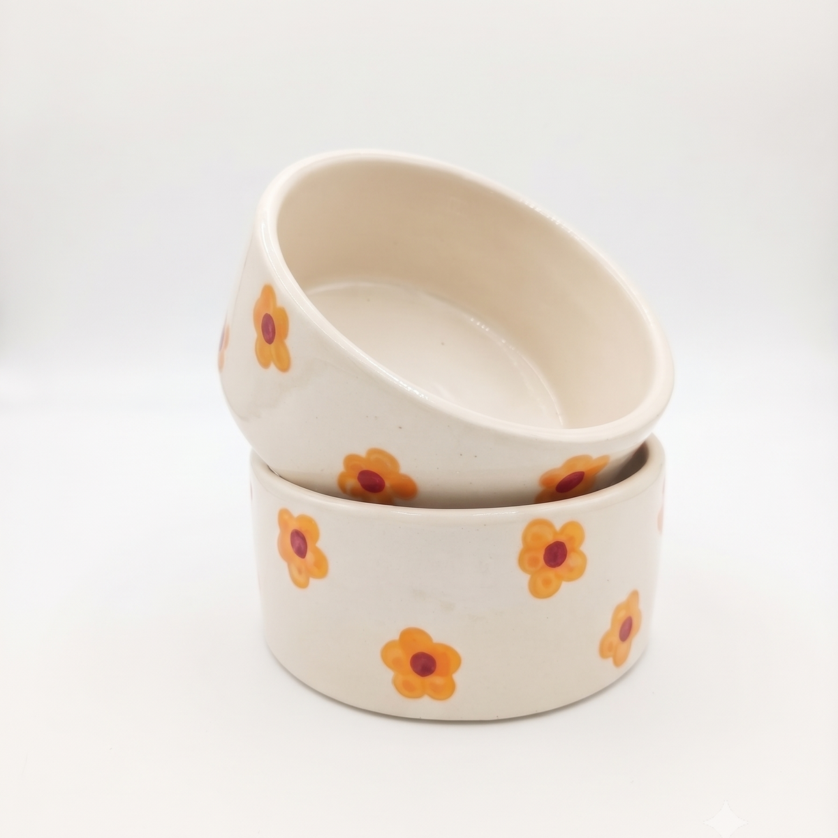 Two stacked ceramic bowls with a warm cream base and a pattern of small orange flowers with dark red centers, set against a seamless white background.