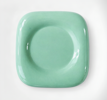 A glossy ceramic square plate with rounded corners and a raised circular center, shown from above on a white background.
