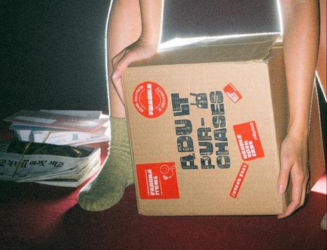 A cardboard box labeled 'ADULT PURCHASER' with red stickers and bold black text, held by a person wearing green socks and denim shorts in a dark setting.
