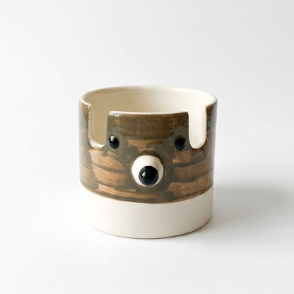A ceramic mug with a sculptural, three-dimensional bear face design. The mug features a glossy brown glazed body with black eyes and a snout, accented by dripping white glaze details and a solid white base, set against a clean white background.
