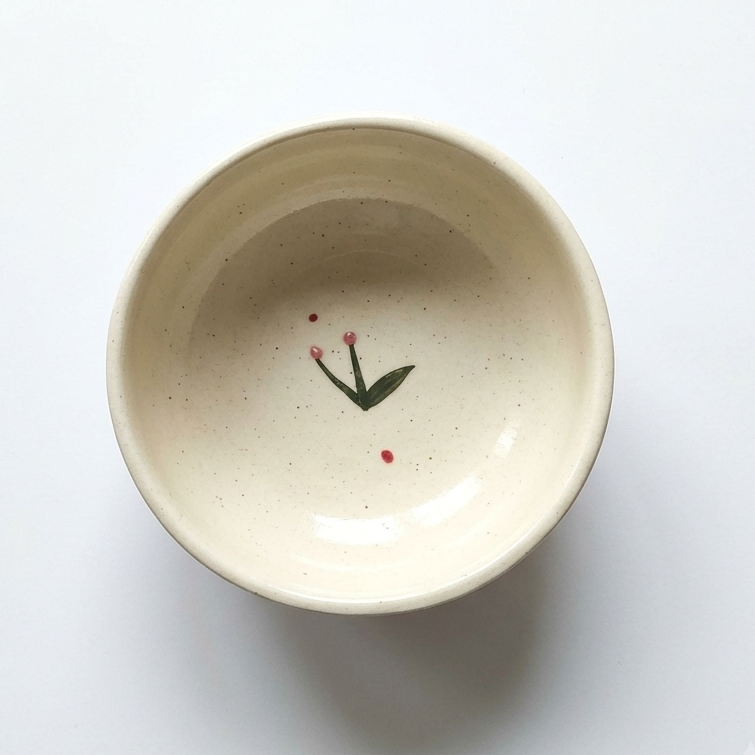 A top-down view of a speckled cream-colored ceramic bowl with a small painted green leaf and red flower motif inside, set against a plain white background.