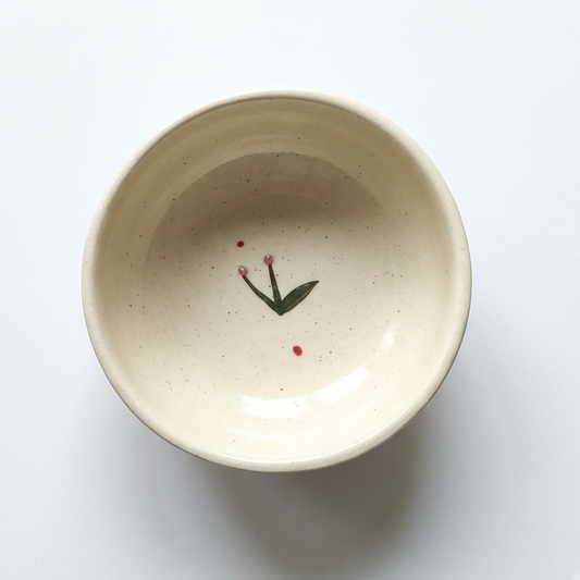 A top-down view of a speckled cream-colored ceramic bowl with a small painted green leaf and red flower motif inside, set against a plain white background.