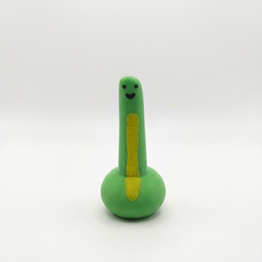 A small green dinosaur-shaped toy with a yellow belly stripe, black facial features, and a round base, photographed against a plain light gray background.