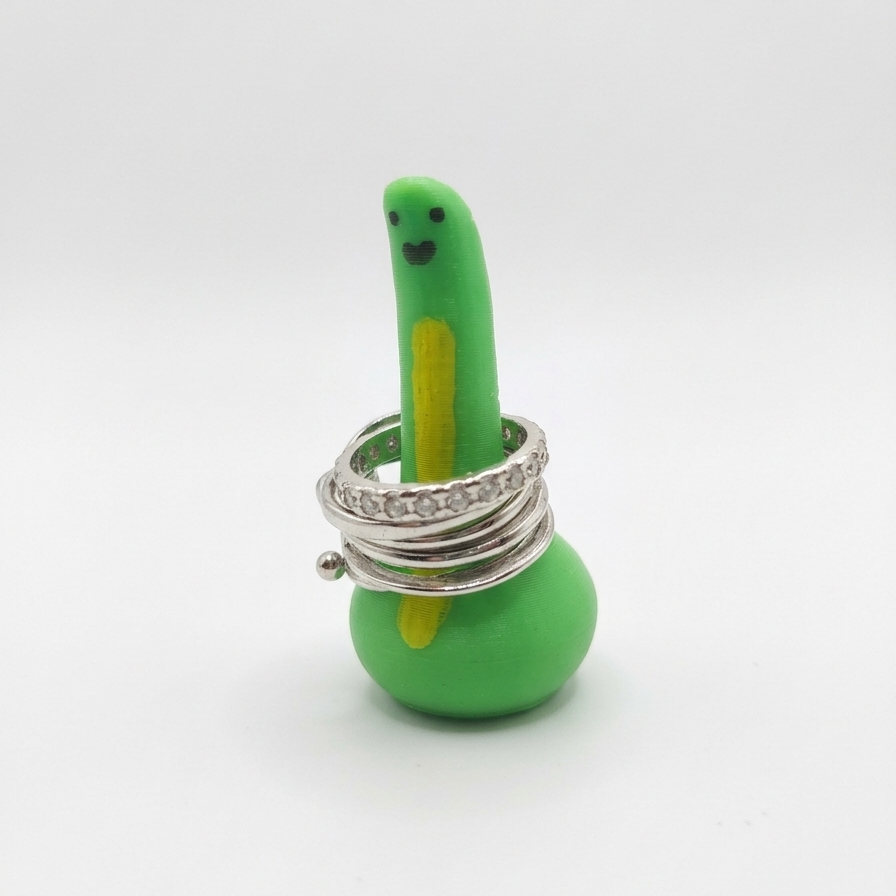 A green 3d printed dinosaur-shaped ring holder with a small smiley face, wrapped with several silver rings, including one with clear rhinestones, on a white background.