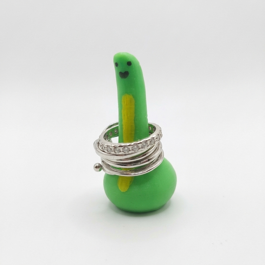 A green 3d printed dinosaur-shaped ring holder with a small smiley face, wrapped with several silver rings, including one with clear rhinestones, on a white background.