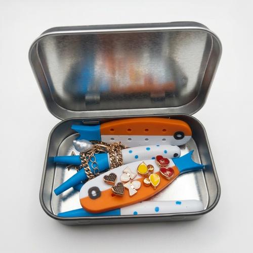 A silver metal tin with its lid open, containing two small fish-shaped tools or accessories in orange, blue, and white, along with several small heart-shaped jewelry charms.