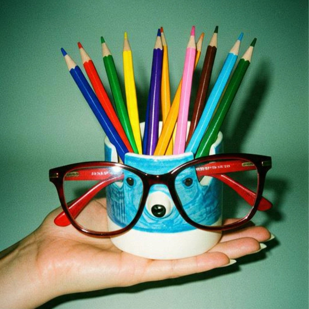 A hand holds a white ceramic pencil cup decorated like a blue polar bear face, filled with colorful pencils, with red-framed glasses resting on top, against a solid teal-green background.