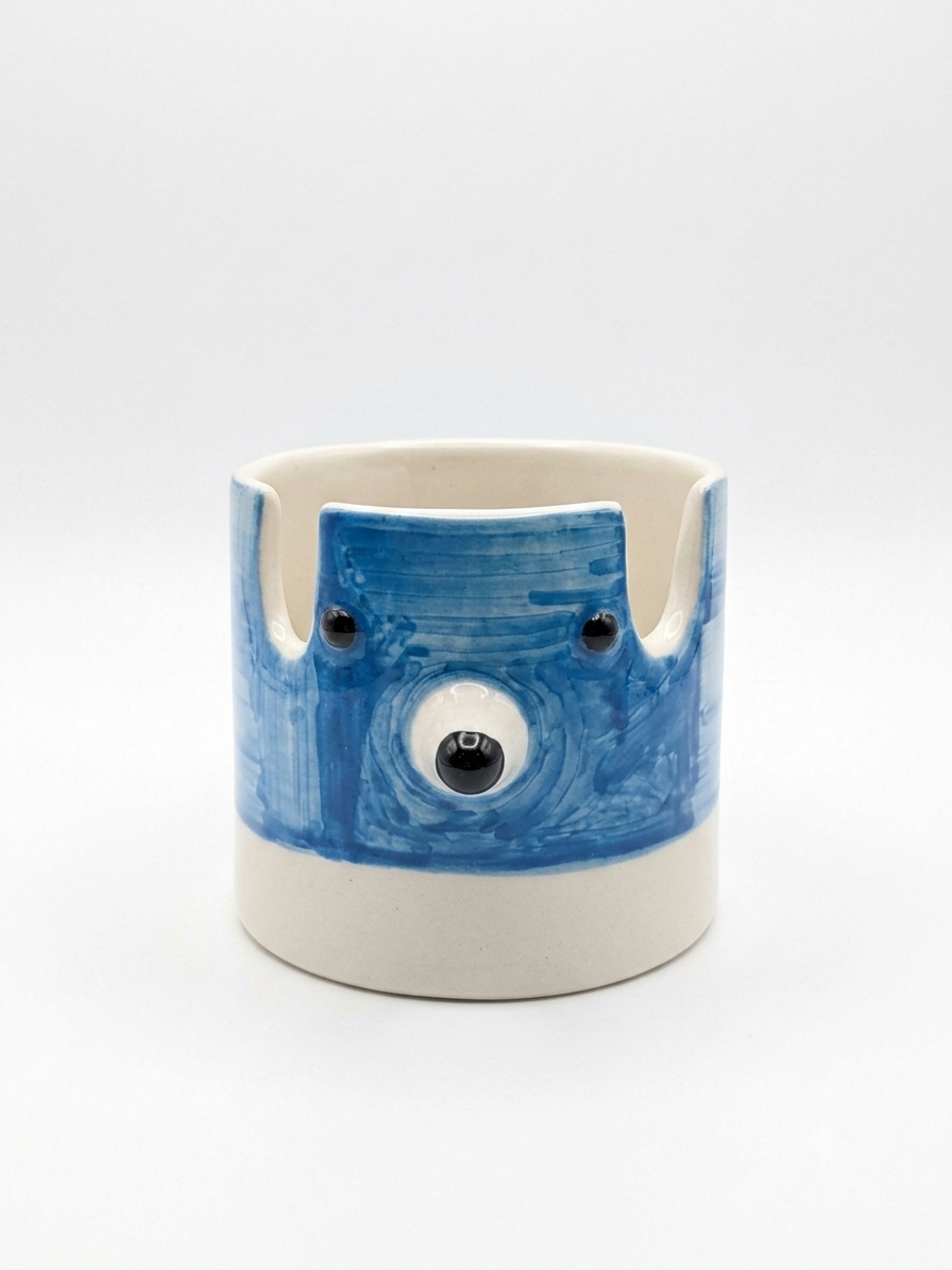 A handmade ceramicpenstand with a blue painted face and black eyes on a cream-colored base, set against a plain white background.