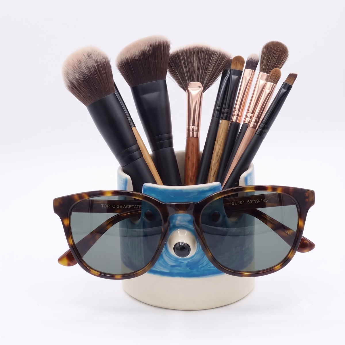 A pair of tortoiseshell acetate sunglasses with dark gray lenses rests on a blue and white ceramic cup filled with makeup brushes. The sunglasses feature the brands 'Tortoise Acetate' and 'Rimowa' printed on the temples.